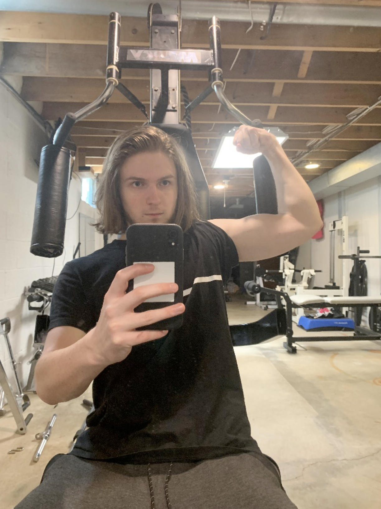 man holding cell phone up (to mirror) of himself in a basement gym, one arm showing muscles